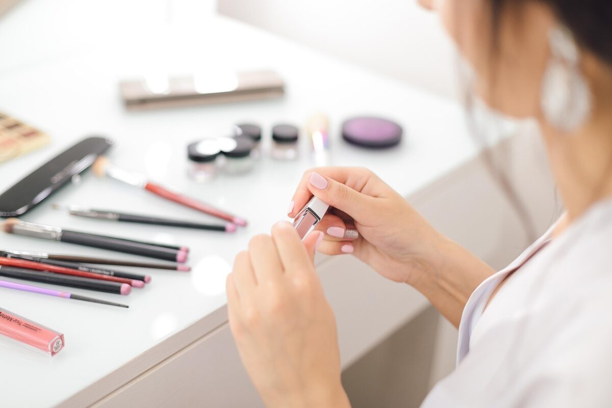 a hand holding a drugstore lipstick swatch next to an upscale lipstick case on a marble countertop